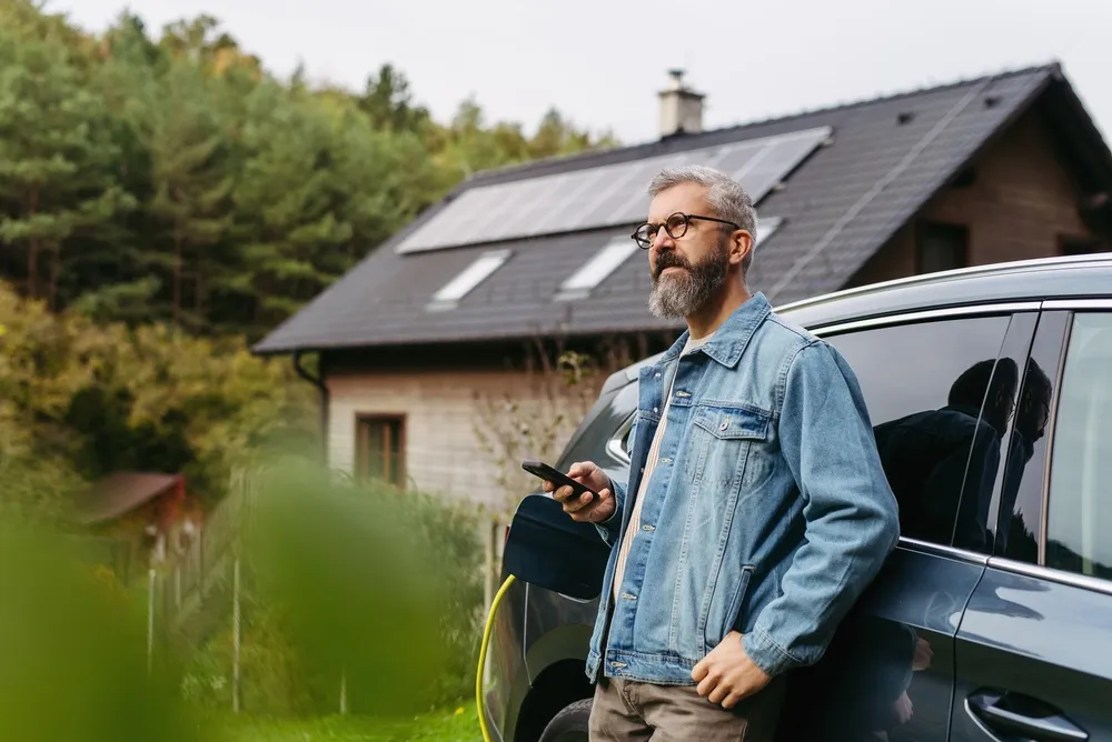 Homme près d'un véhicule électrique en recharge devant une maison équipée de panneaux solaires.